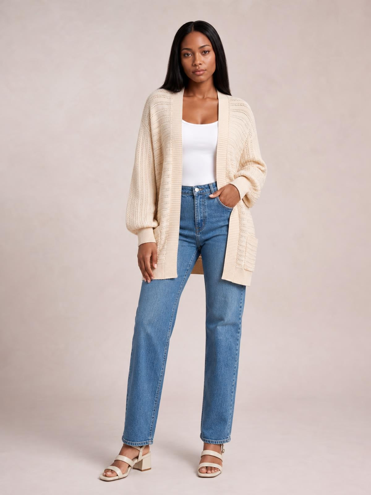 A model stands in a cream open-front sweater cardigan with longline silhouette, ribbed trim, textured knit construction, and patch pockets over a white top and jeans.