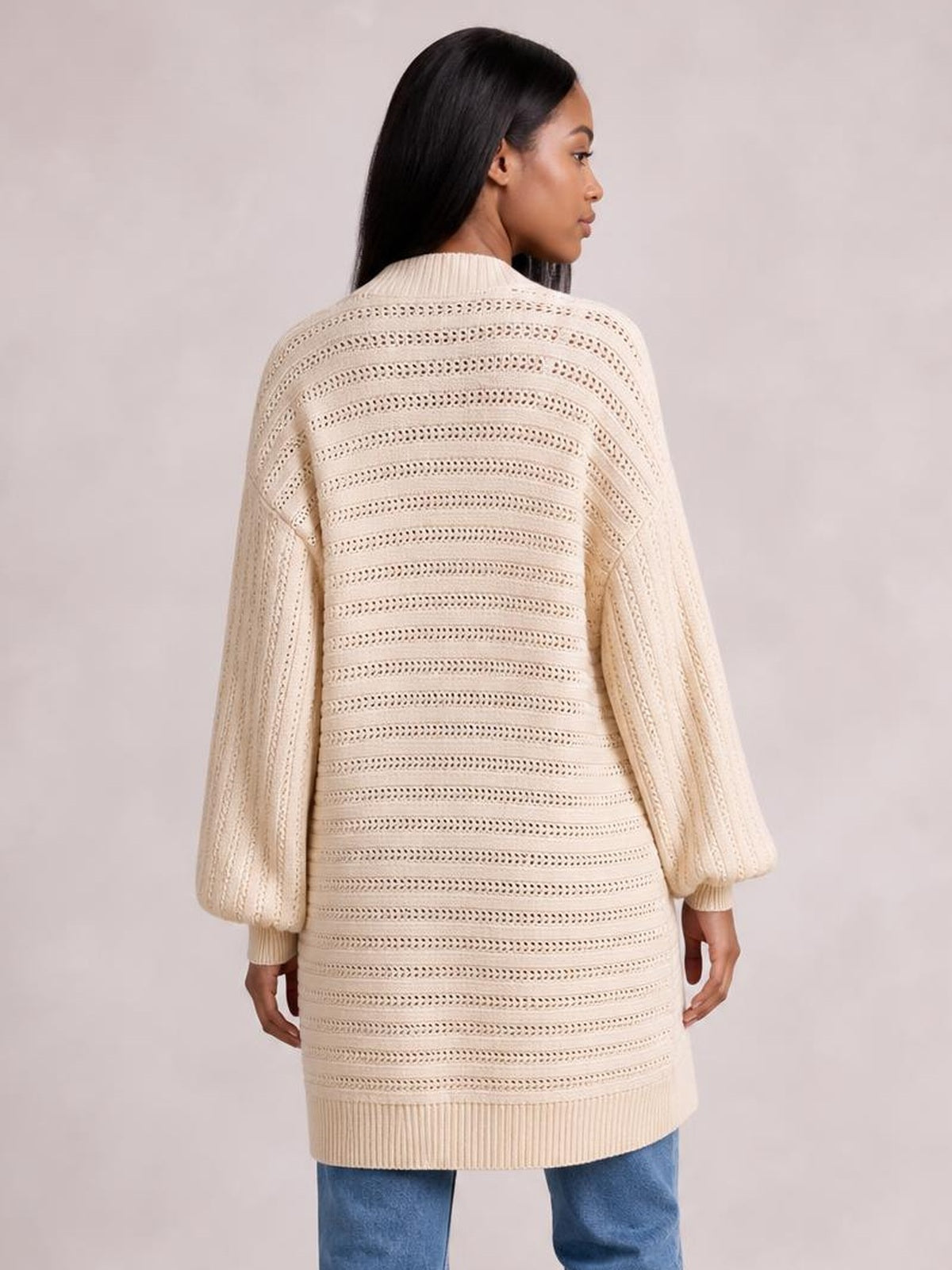 A model shown from behind wears a cream open-front sweater cardigan with an open-knit striped texture, long sleeves, and a relaxed long silhouette.