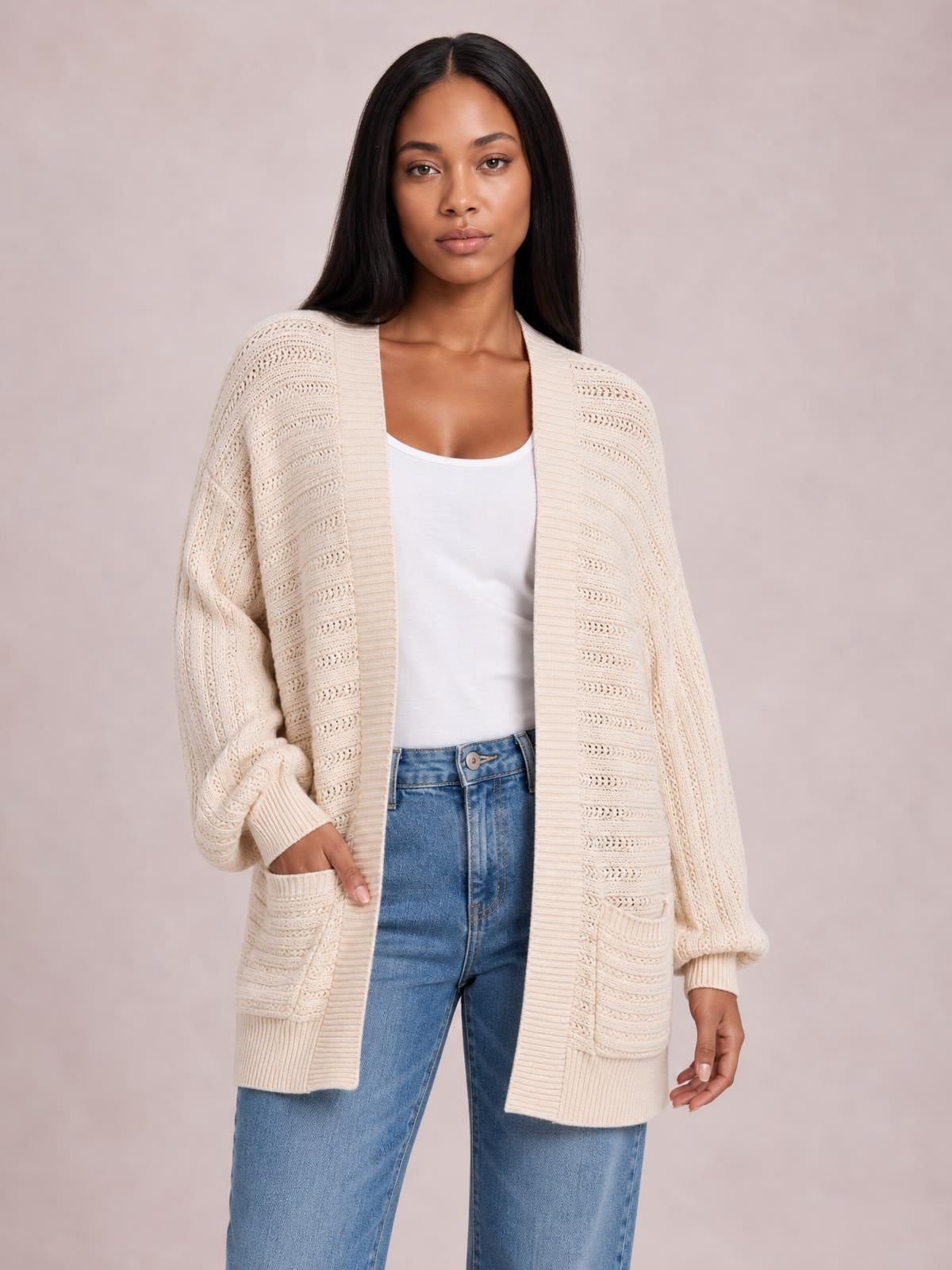 Model wearing an ivory open front sweater cardigan with a relaxed long silhouette, textured knit, ribbed trim, and patch pockets, one hand tucked inside.