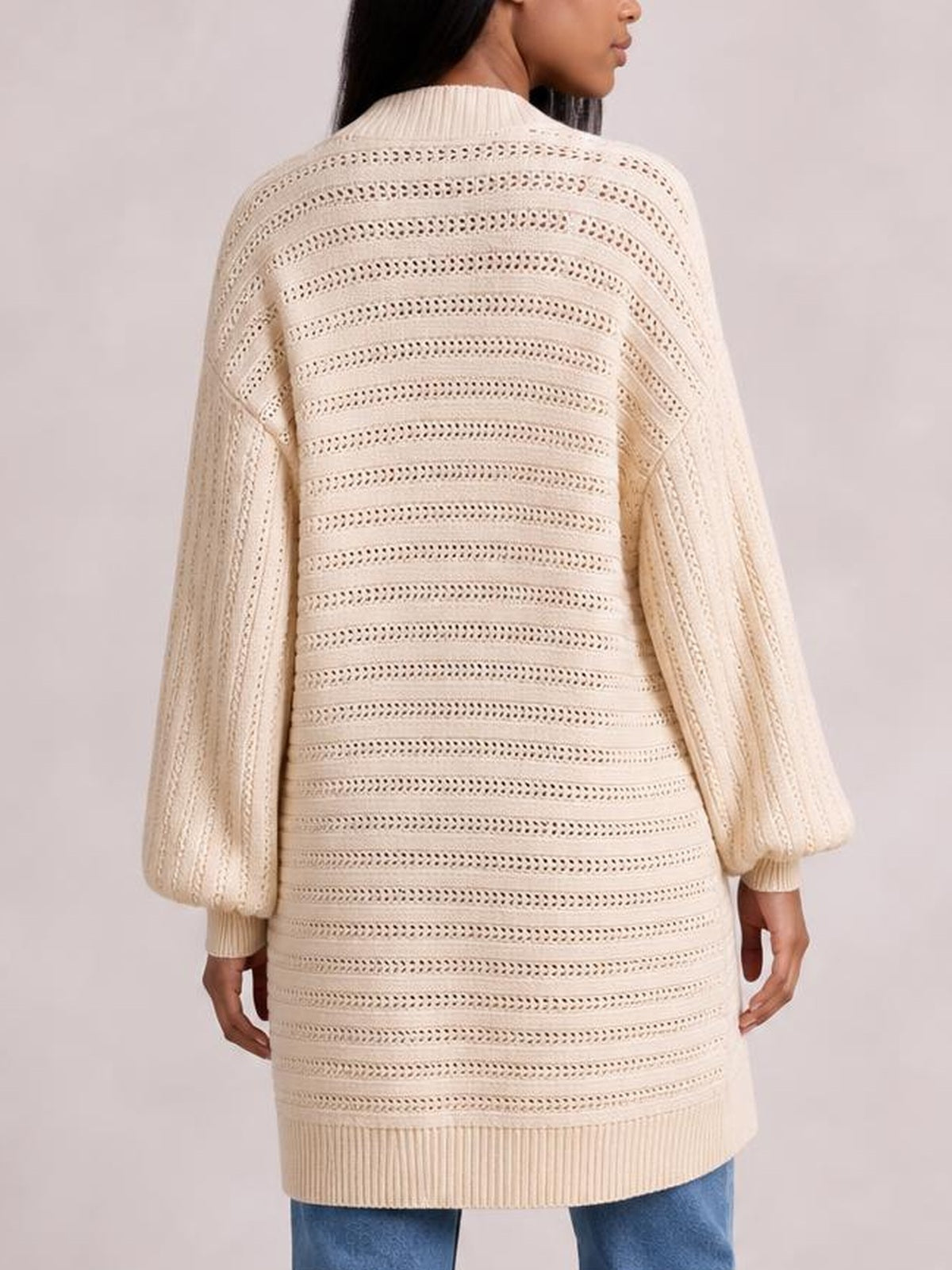 A model stands in a cream sweater cardigan, shown from the back with a relaxed long silhouette, openwork horizontal knit, and ribbed collar, cuffs, and hem.