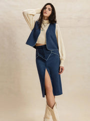 Model poses with one hand behind her head in a peacock blue open-front vest and buttoned midi skirt set with contrast piping and a front slit.