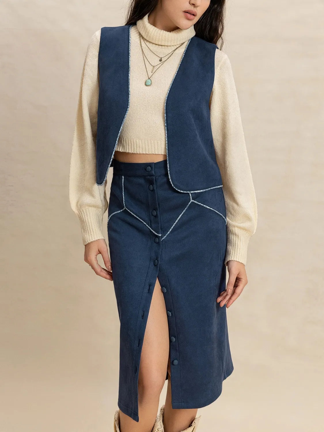 Model standing in a peacock blue open-front vest and buttoned midi skirt set with curved edges, contrast trim, and a front slit over a cream sweater.