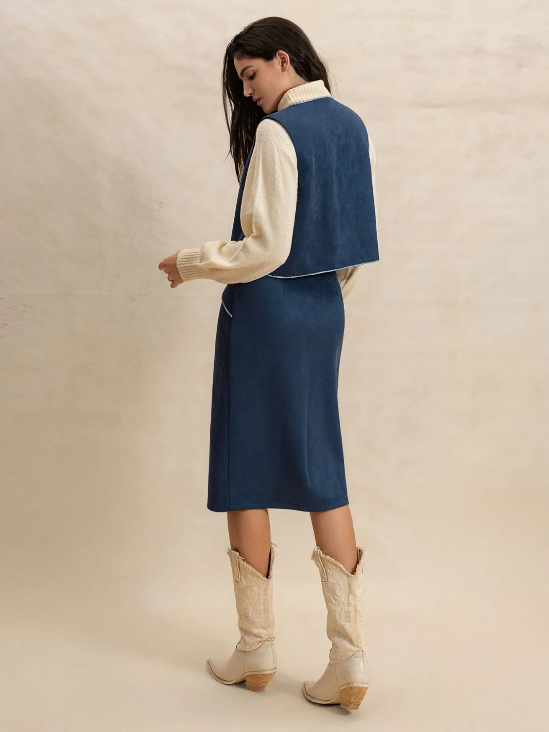 Model turned away shows a peacock blue open-front vest with softly curved edges over a cream knit and matching straight midi skirt for a polished layered silhouette.