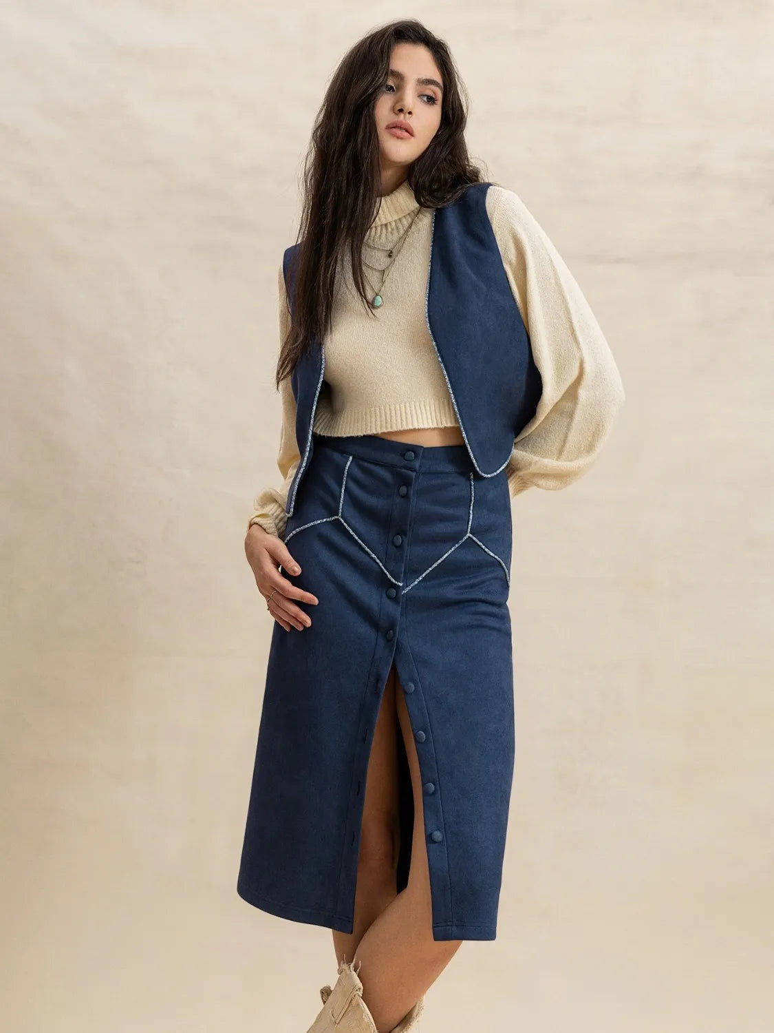 Model poses with one hand on her hip in a peacock blue open-front vest and buttoned midi skirt set over a cream knit, creating a polished layered silhouette.