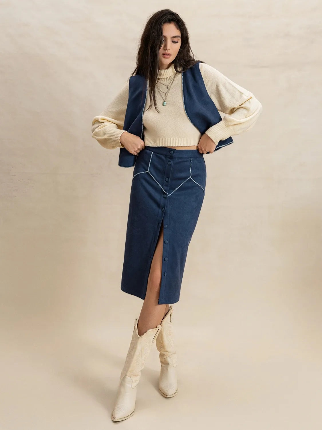 A model poses with hands at her waist in a peacock blue open-front sleeveless vest and matching cropped midi skirt over a cream knit, with front buttons and a slit.