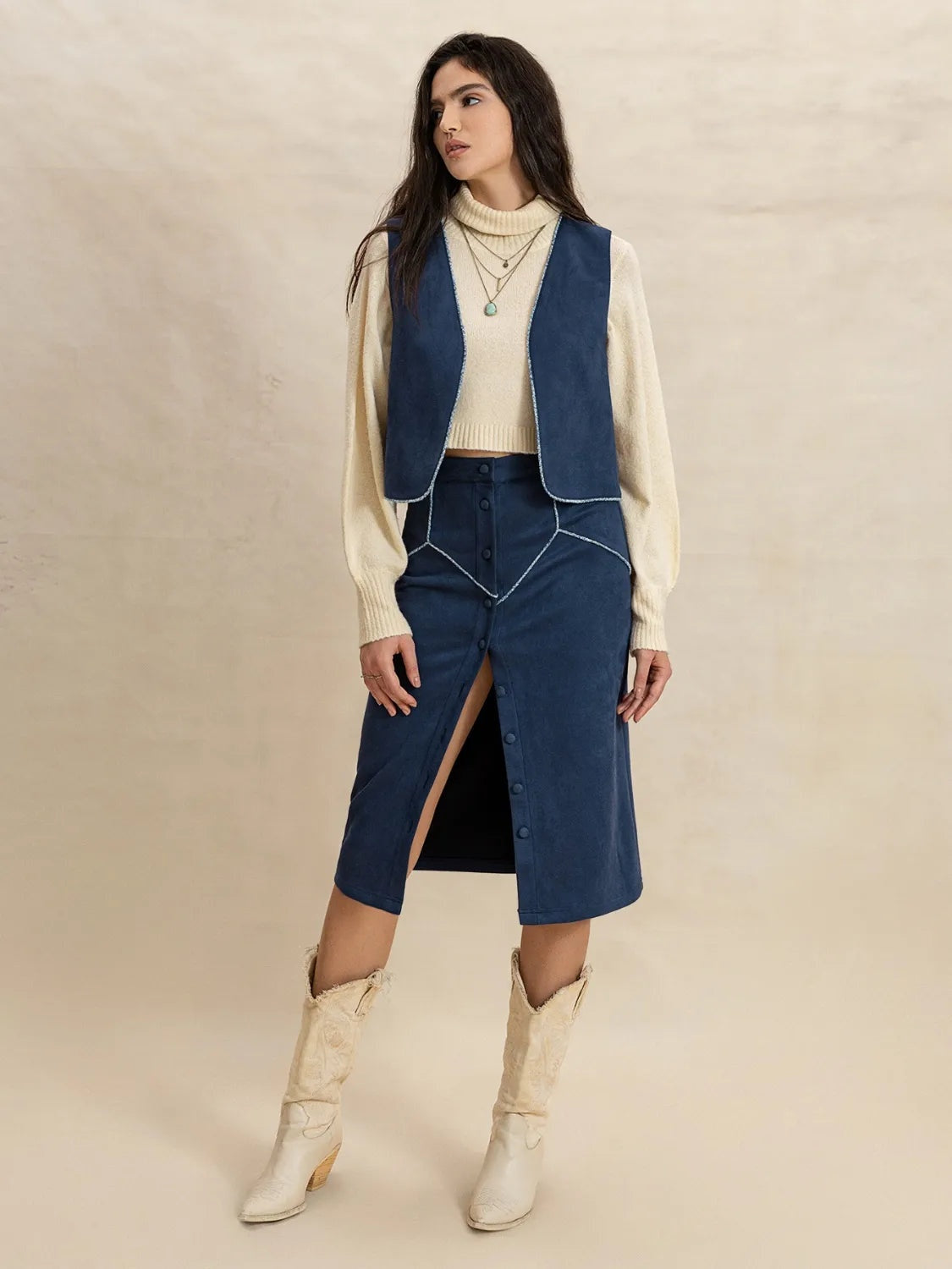 Model stands in a peacock blue open-front vest and buttoned midi skirt set, featuring a cropped silhouette, curved edges, and front slit.