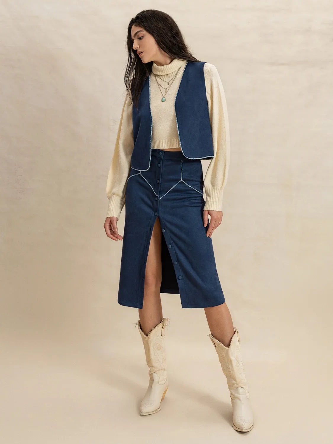A model poses in a Peacock Blue open-front vest with curved edges and a matching buttoned midi skirt featuring a front opening over a cream knit.