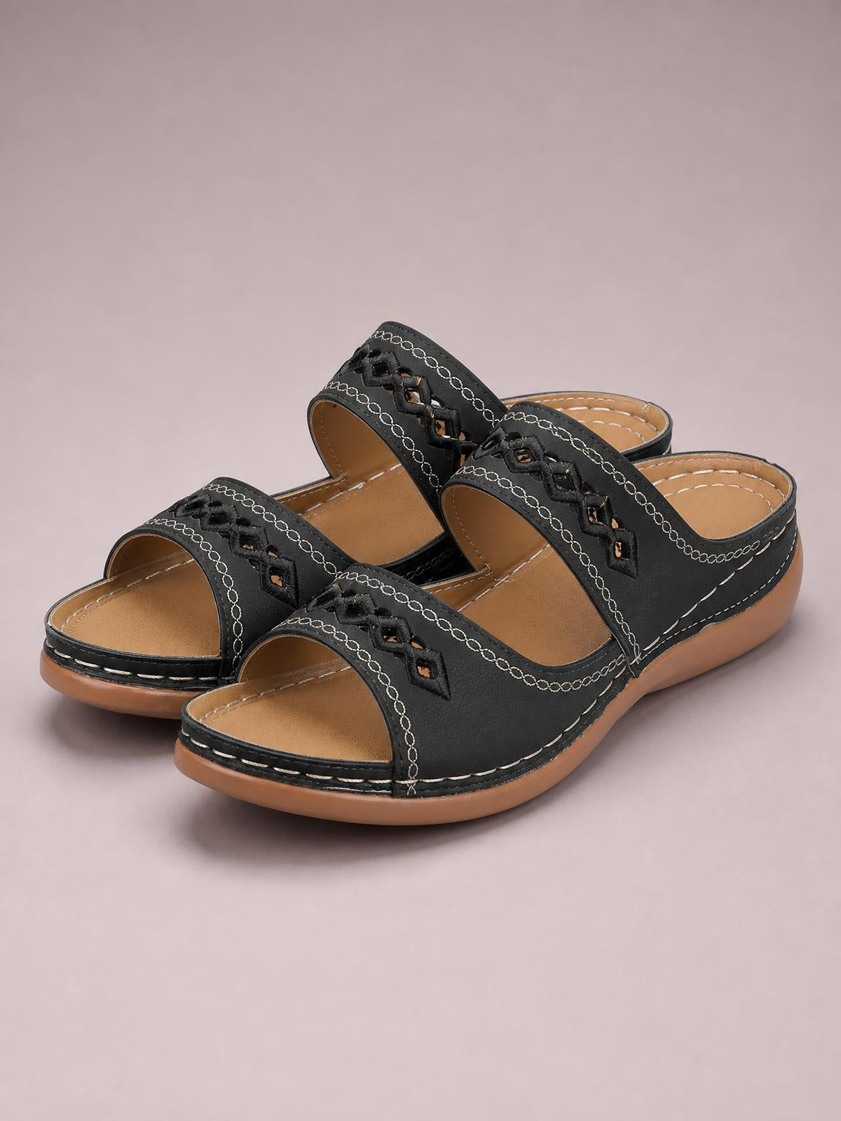 Black open-toe flat sandals in PU leather feature woven front straps, decorative white stitching, tan footbeds, and sturdy rubber soles.