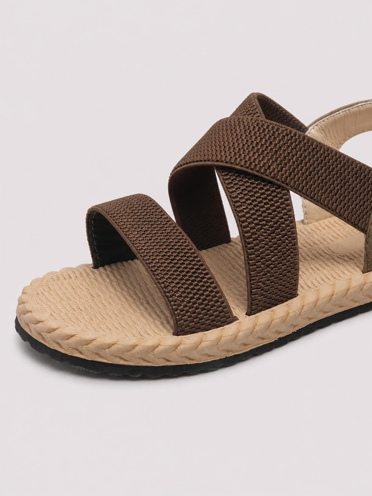 Brown open toe flat sandals feature wide woven straps, a textured beige footbed, braided edge detail, and a black rubber outsole.