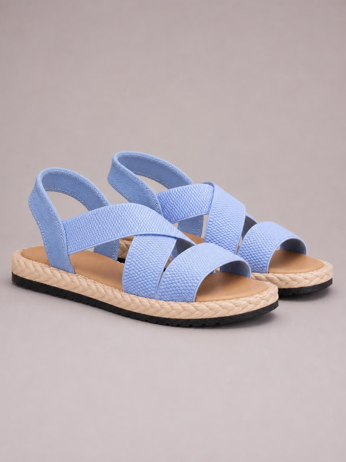 Light blue open toe flat sandals feature wide woven straps, adjustable heel straps, cushioned footbeds, braided midsoles, and black rubber outsoles.