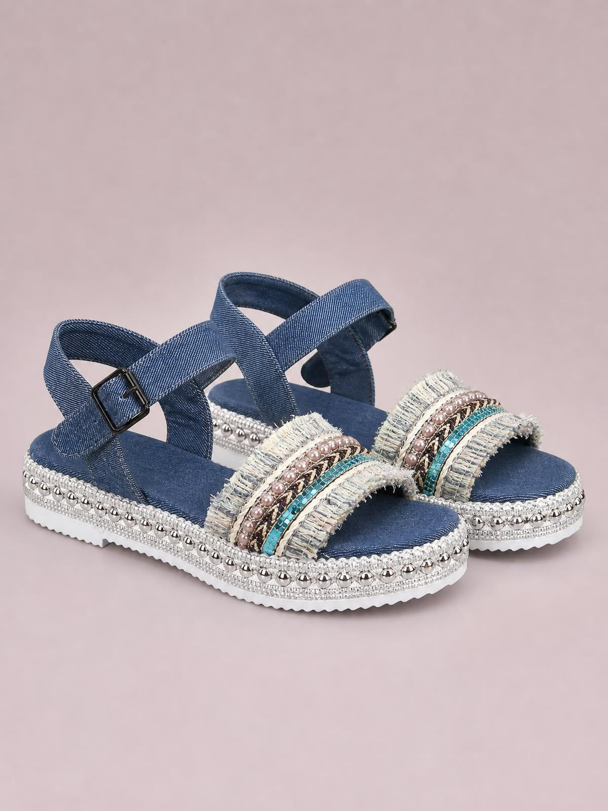 Blue denim open toe platform sandals feature embellished fringed straps, ankle buckles, studded trim, and a white rubber sole with a flat silhouette.