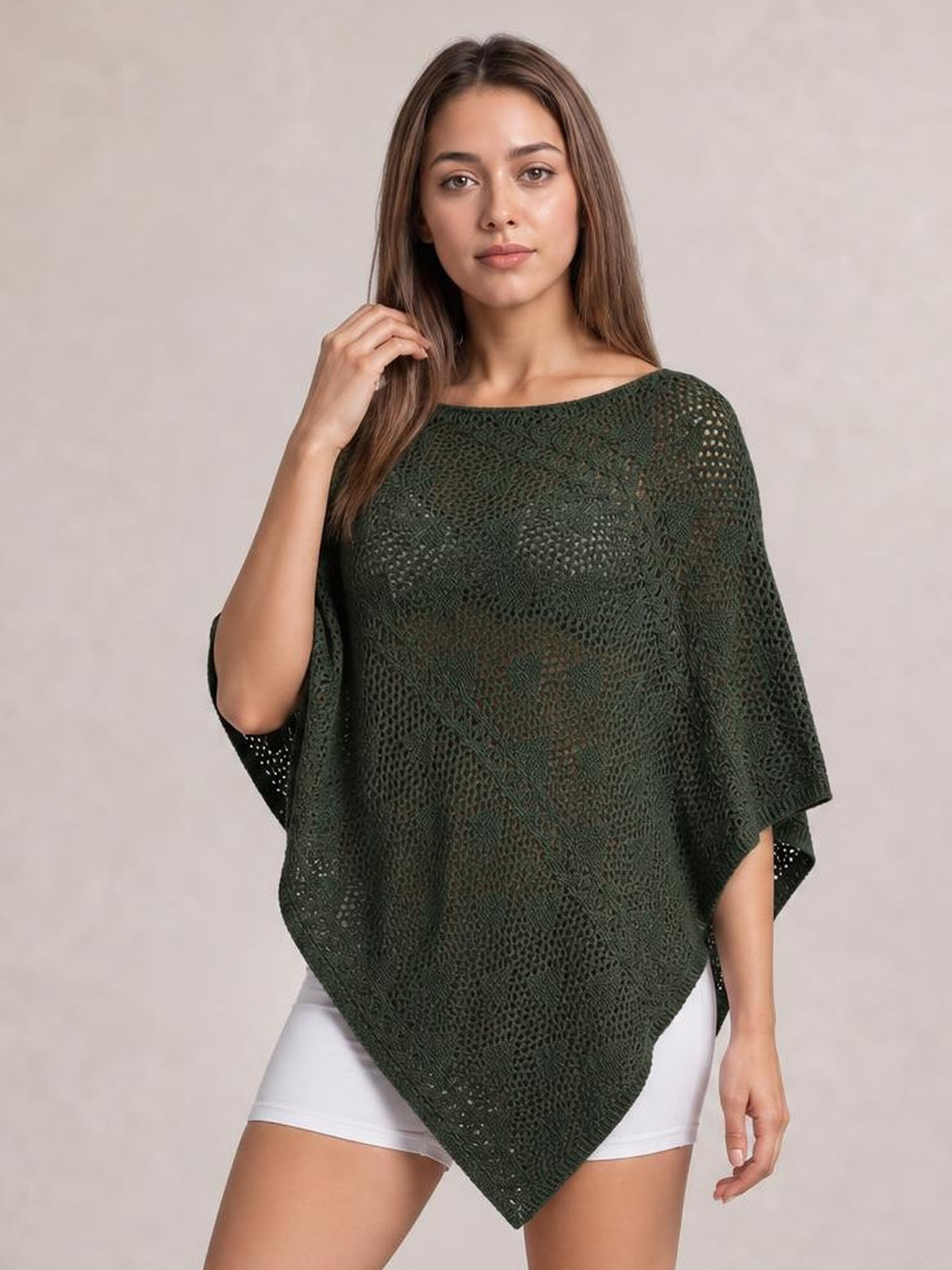 Model poses in a dark green openwork boat-neck shawl cover up with a loose poncho silhouette, draped wide sleeves, and a pointed hem over white shorts.