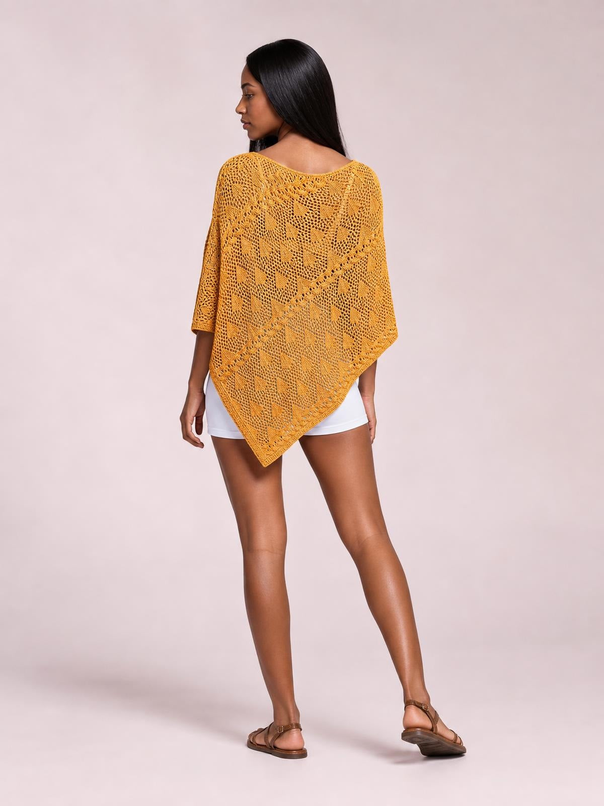 Model in a back-facing pose wears a mustard openwork boat neck shawl cover up with a loose poncho silhouette, flowing pointed hem, and three-quarter sleeves.