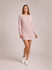 Model wears a pale pink openwork boat neck sweater dress with long sleeves, ribbed cuffs and hem, a fitted mini silhouette, and one leg crossed.