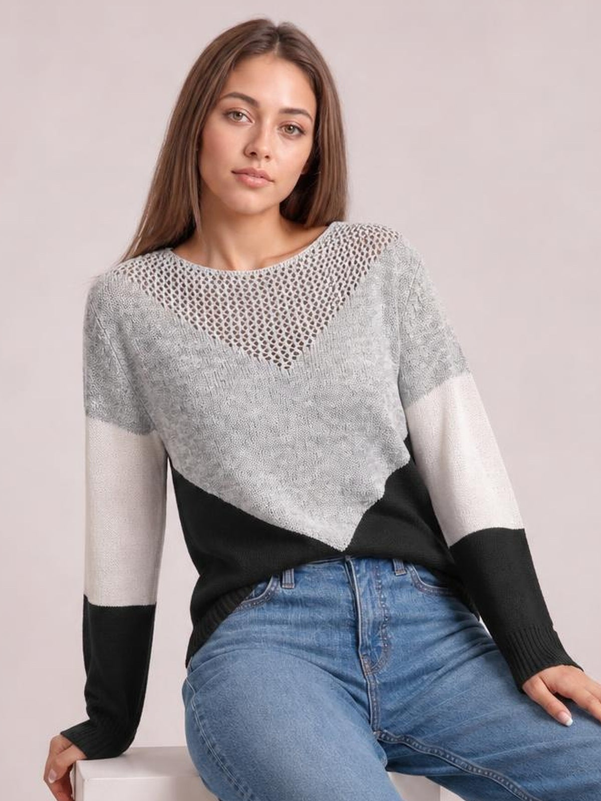 Model seated in a gray and black color block long sleeve sweater with an openwork neckline, relaxed silhouette, and contrasting ribbed cuffs.