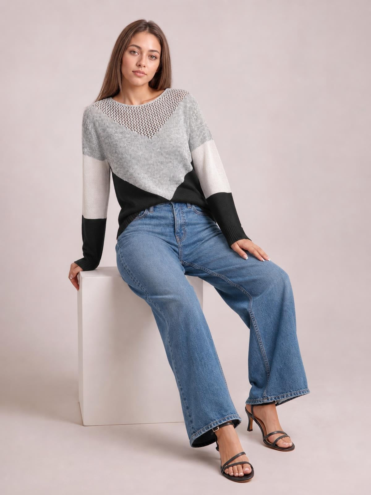 Model seated in a relaxed pose wears a gray, white, and black color block sweater with long sleeves, an openwork neckline, and a relaxed silhouette.