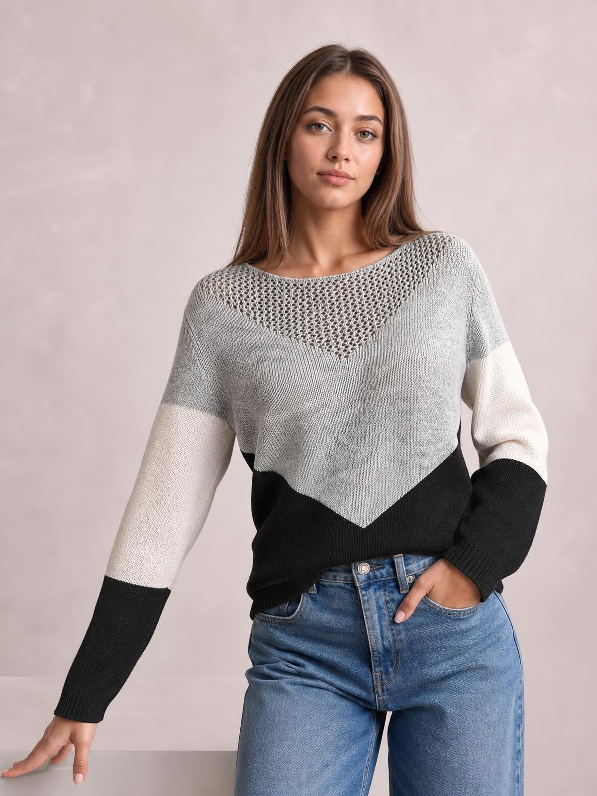 Model wearing a gray, white, and black color block long sleeve sweater with openwork neckline, relaxed silhouette, and ribbed cuffs, posing with one hand in her jeans pocket.