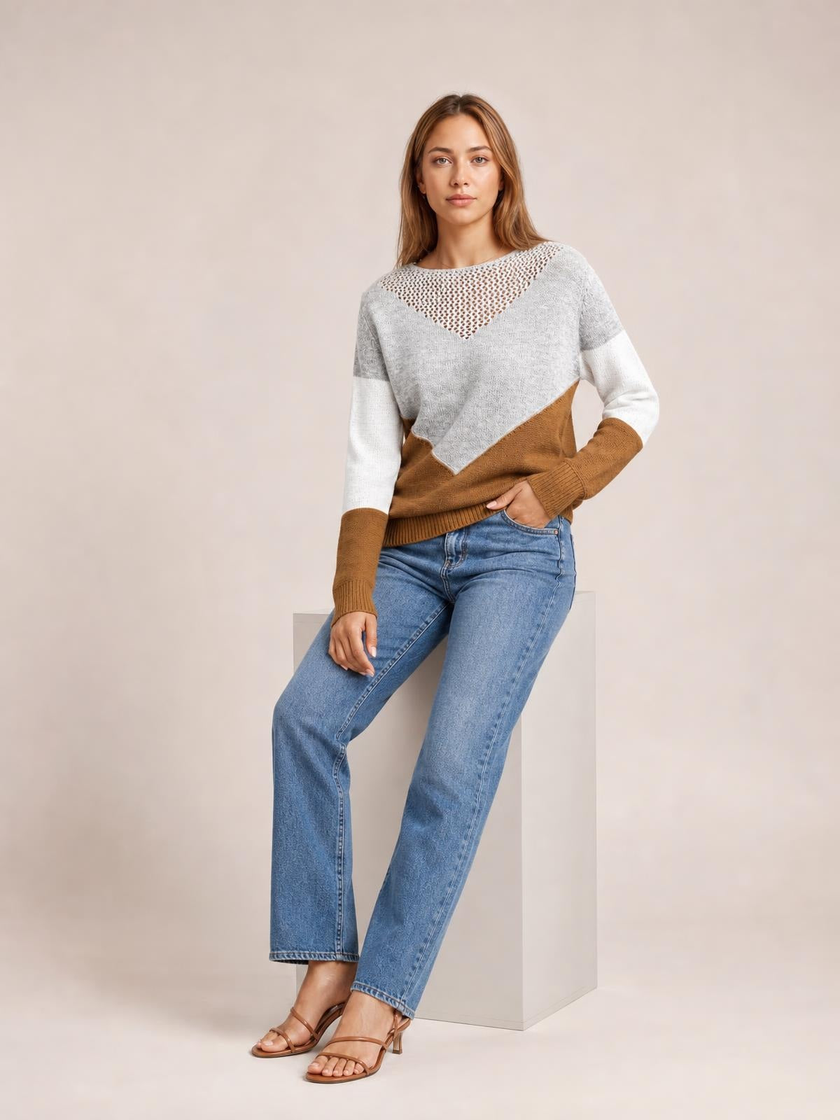 A seated model wears a relaxed color block long sleeve sweater in gray, white, and brown with an openwork triangular neckline and ribbed hem.