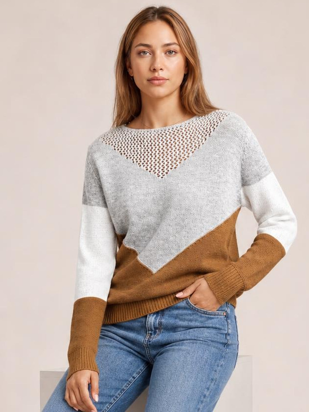 A woman wears a gray, white, and brown color-block sweater with long sleeves, openwork neckline, ribbed cuffs and hem, posing with one hand in her jeans pocket.