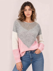Model wears a relaxed color block sweater with gray, pink, and cream panels, long sleeves, ribbed cuffs, and an openwork neckline while posing with one hand in a pocket.