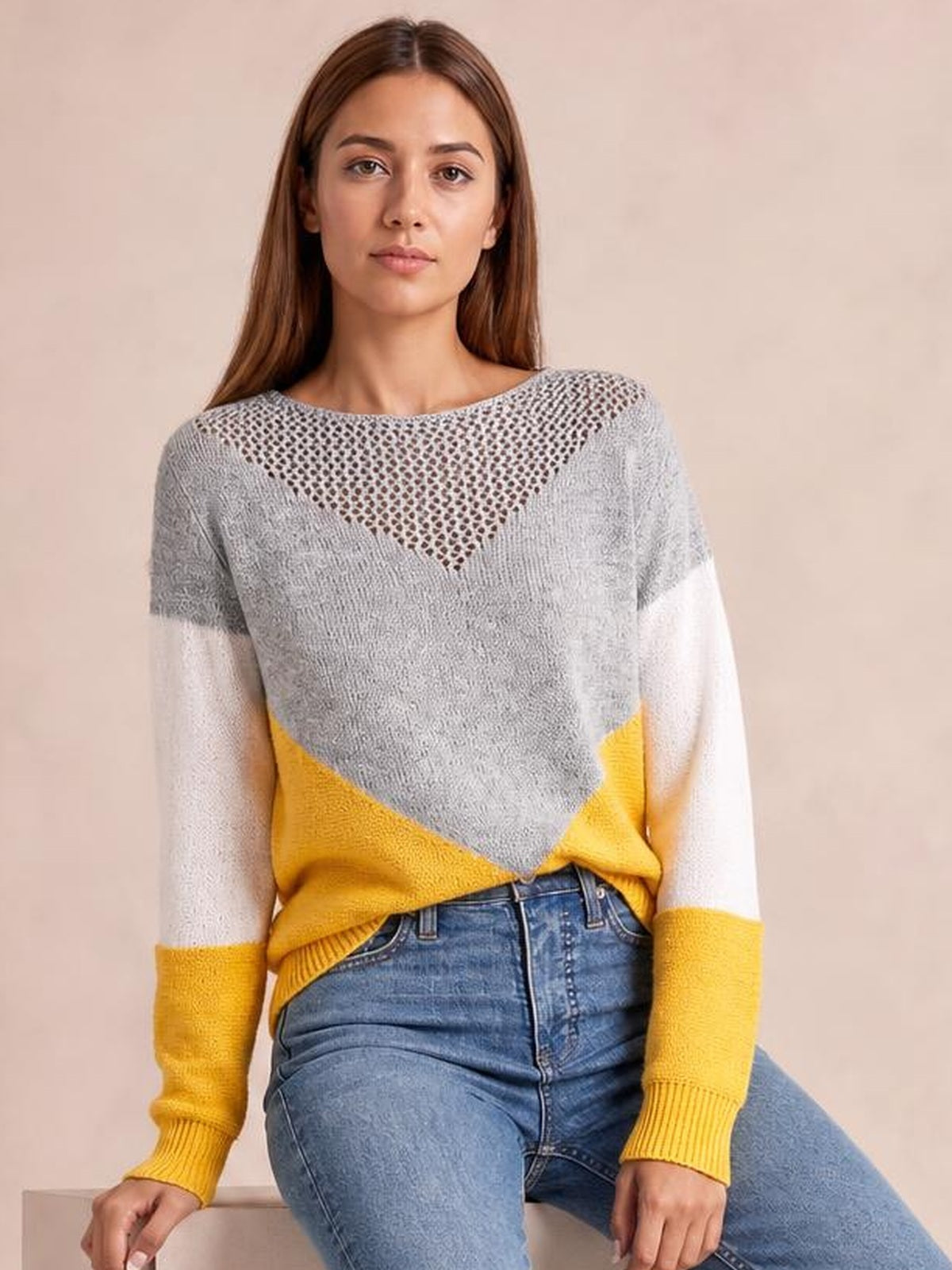 Model seated and facing forward in a long-sleeve openwork color block sweater with gray, white, and yellow panels and a relaxed silhouette.