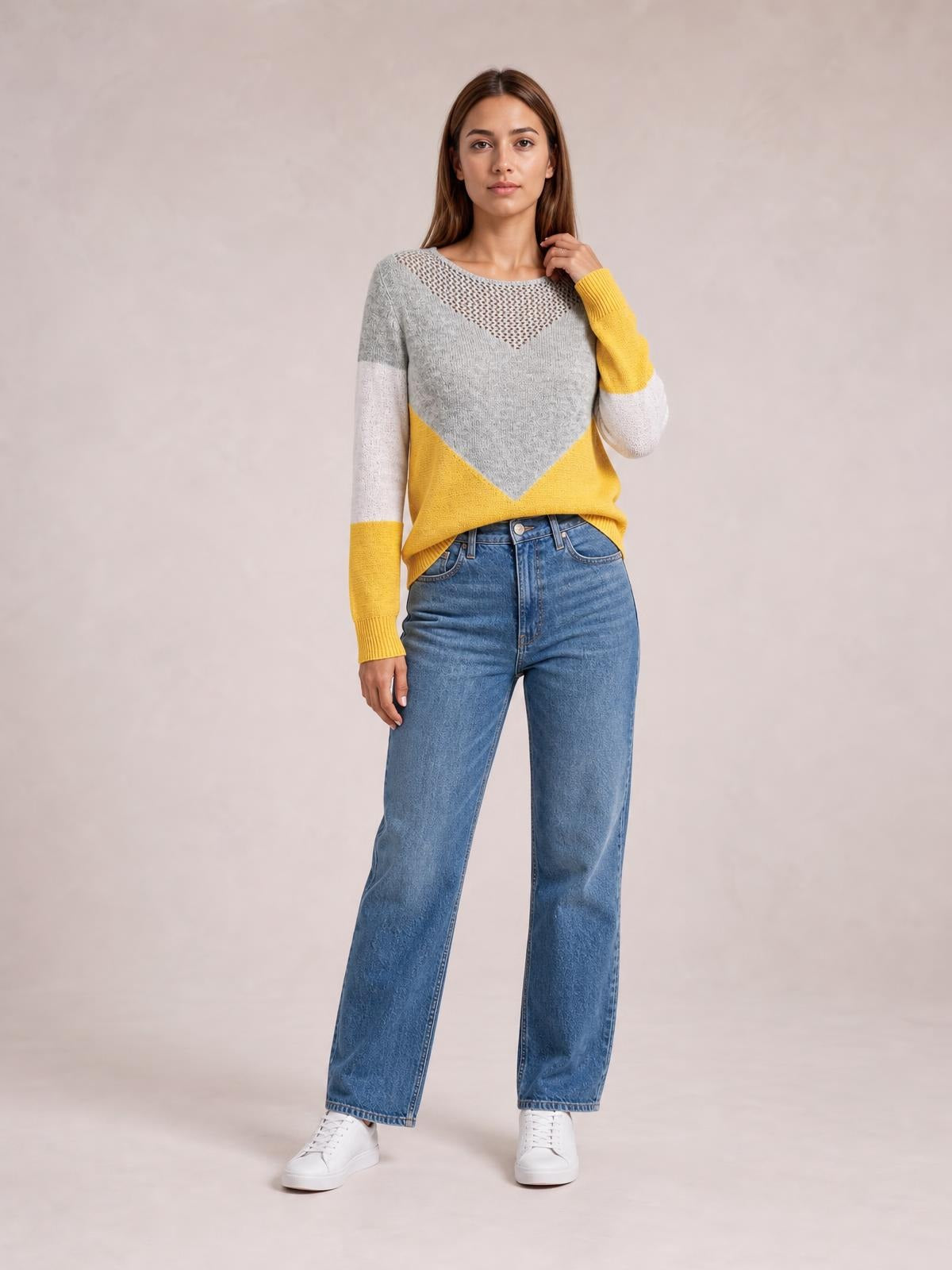 Model wearing a gray, mustard yellow, and white color block long sleeve sweater with an openwork neckline and relaxed silhouette, standing with one hand near her hair.