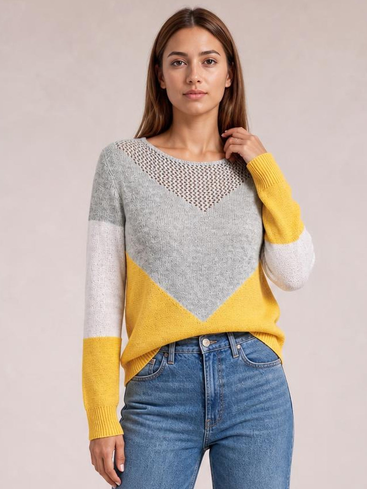 Model wearing a relaxed color block sweater with gray and yellow panels, long sleeves, ribbed cuffs, and an openwork neckline while touching her hair.