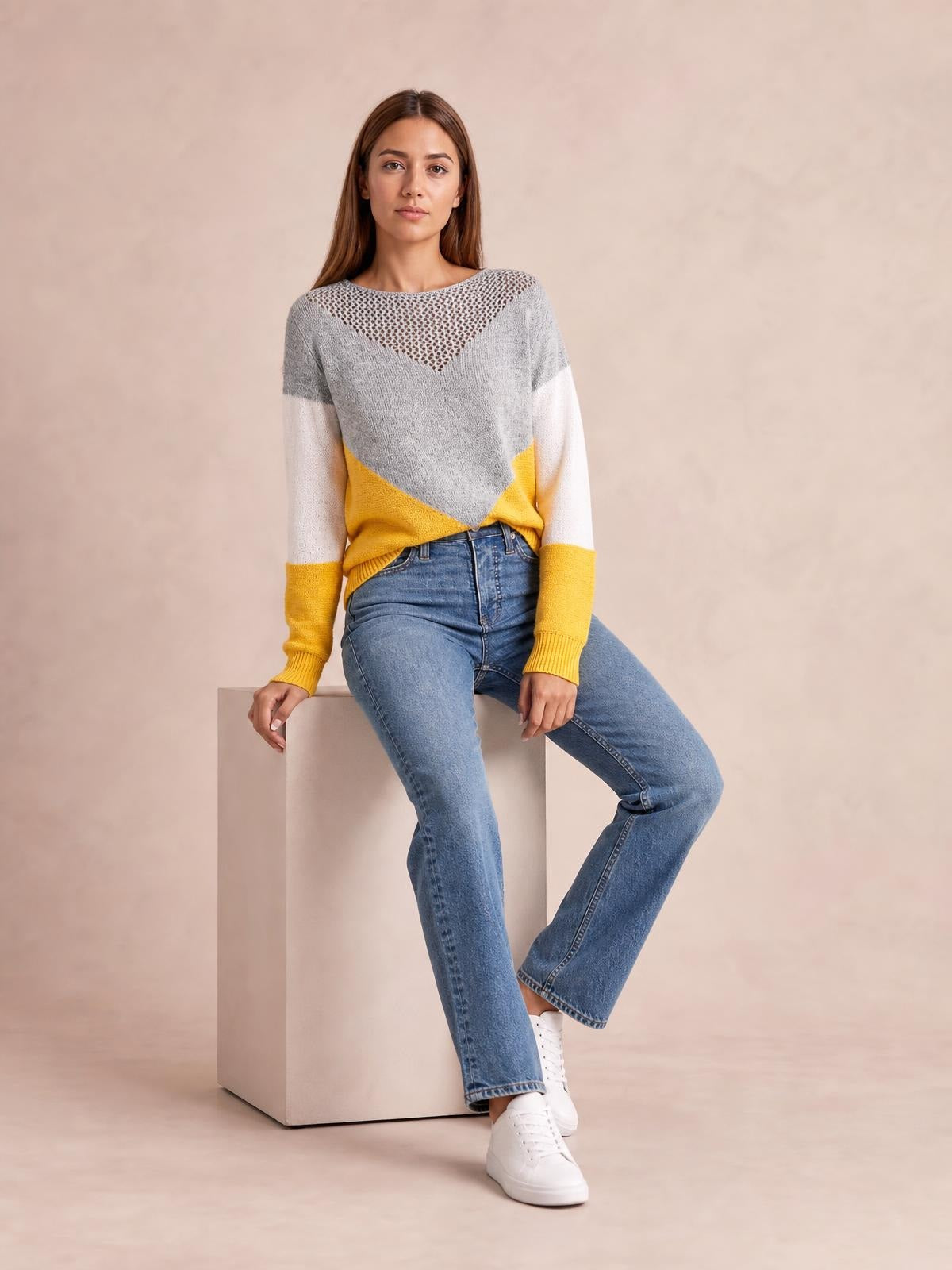Model seated in a relaxed pose wears a gray, white, and mustard color block long sleeve sweater with an openwork V-shaped neckline and relaxed silhouette.