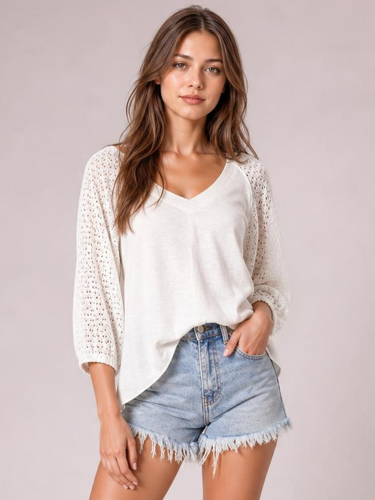 A model wears a white oversized V-neck top with openwork three-quarter sleeves, exposed seams, and a curved high-low hem over denim shorts.