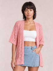 Model wearing a pink openwork open-front cardigan with half sleeves, ribbed edges, and a relaxed long silhouette over a white cropped top.