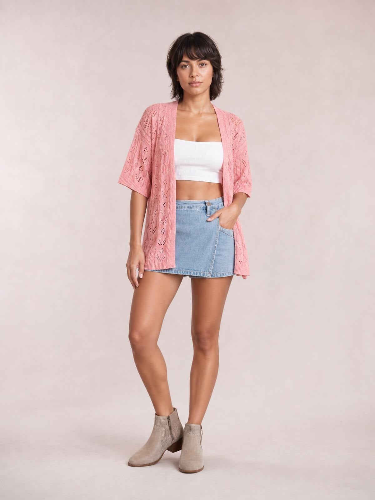 Model poses in a pink openwork open-front cardigan with half sleeves, a relaxed long silhouette, and patterned knit construction over a white crop top.
