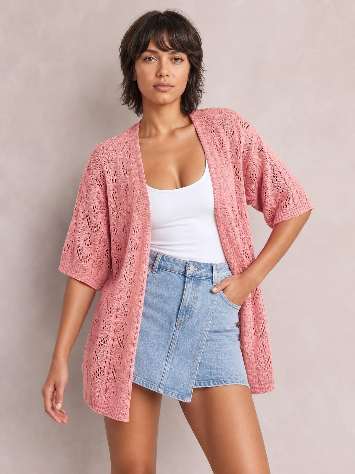 Model wearing a dusty pink openwork open-front cardigan with loose half sleeves and a long relaxed silhouette over a white tank, posing with one hand in her pocket.