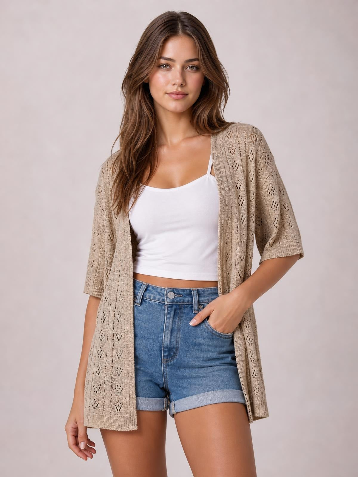 Model poses in a beige openwork open-front cardigan with half sleeves and a relaxed long silhouette over a white camisole and denim shorts.