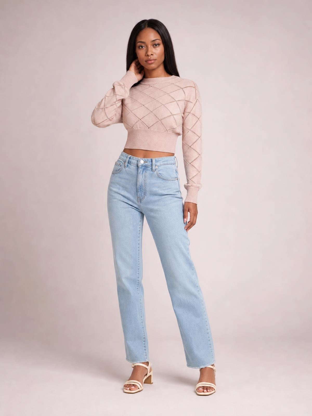 Model wearing a blush pink openwork plaid round-neck cropped sweater with long sleeves, ribbed cuffs and hem, posing with one hand near her neck.