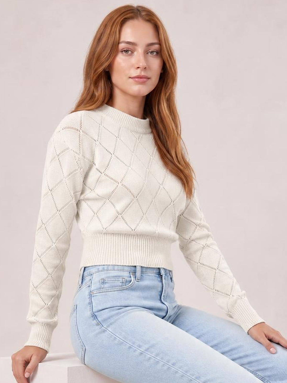 Seated model wears an ivory openwork plaid cropped sweater with a round neck, long sleeves, ribbed cuffs and hem, and a fitted silhouette.