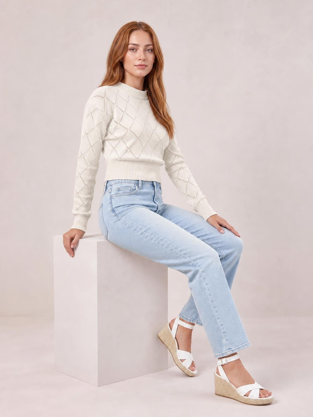 A seated model wears an ivory cropped sweater with a round ribbed neck, long sleeves, ribbed cuffs and hem, and an openwork diamond pattern.