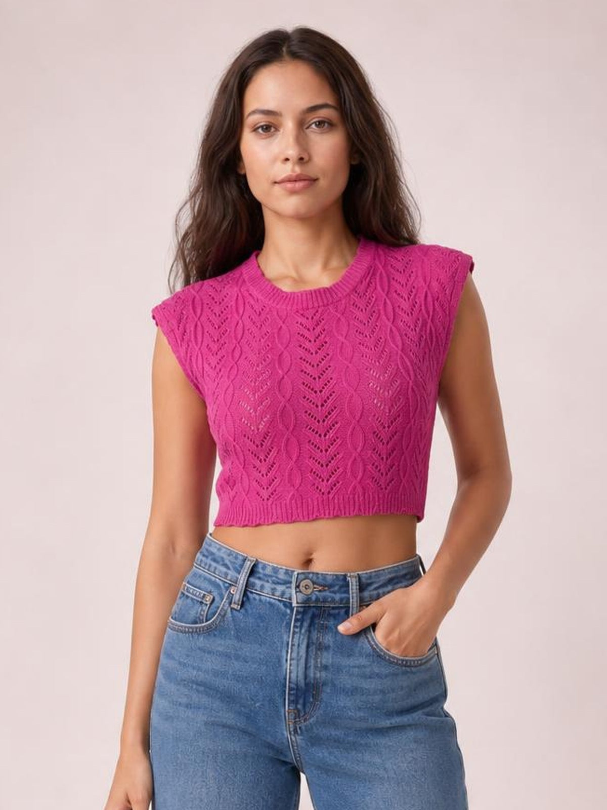 Model wears a bright pink openwork knit top with a round neck, cap sleeves, ribbed trim, and a cropped silhouette, posing with one hand in her jeans pocket.