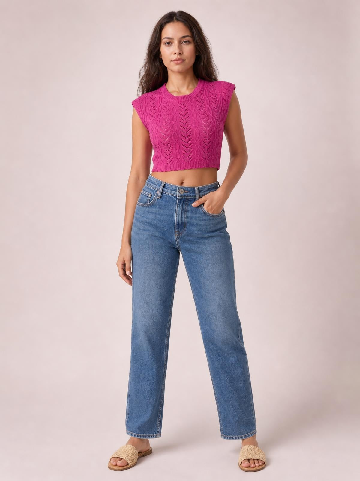 Model wears a fuchsia openwork round neck knit top with cap sleeves, a ribbed hem, and a cropped silhouette, posing with one hand in her jeans pocket.