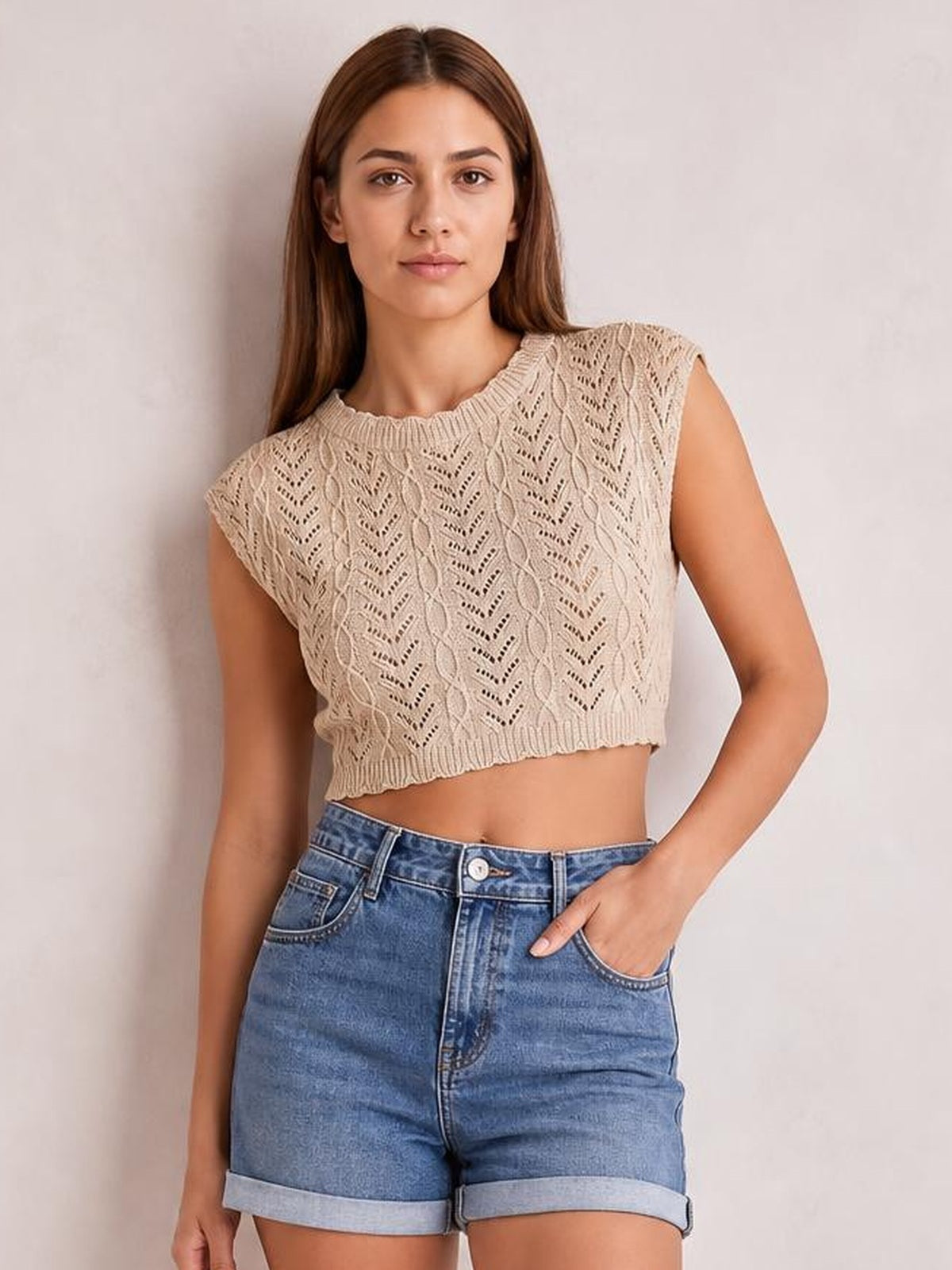 Model wears a beige openwork round neck cap sleeve knit top with a cropped silhouette, ribbed trim, and textured patterned construction, posing with one hand in her jeans pocket.