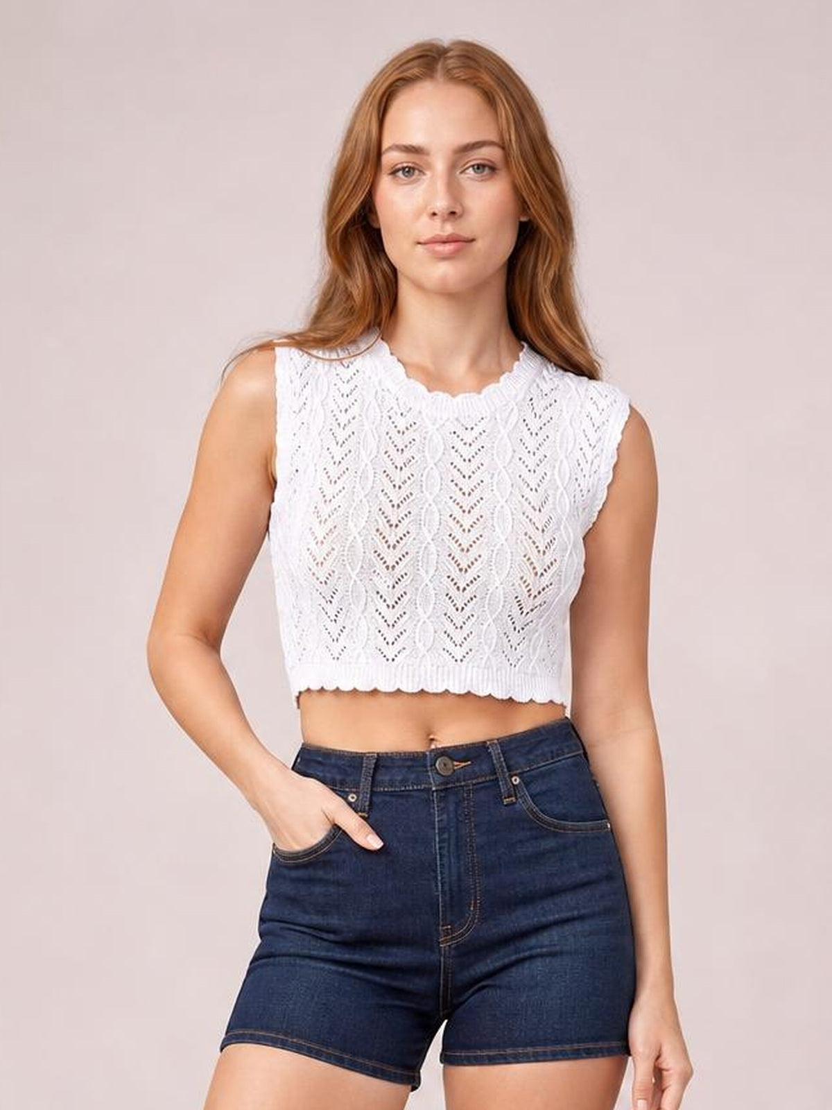Model poses with one hand in her pocket wearing a white openwork knit top with a round neck, cap sleeves, scalloped edges, and a cropped silhouette.