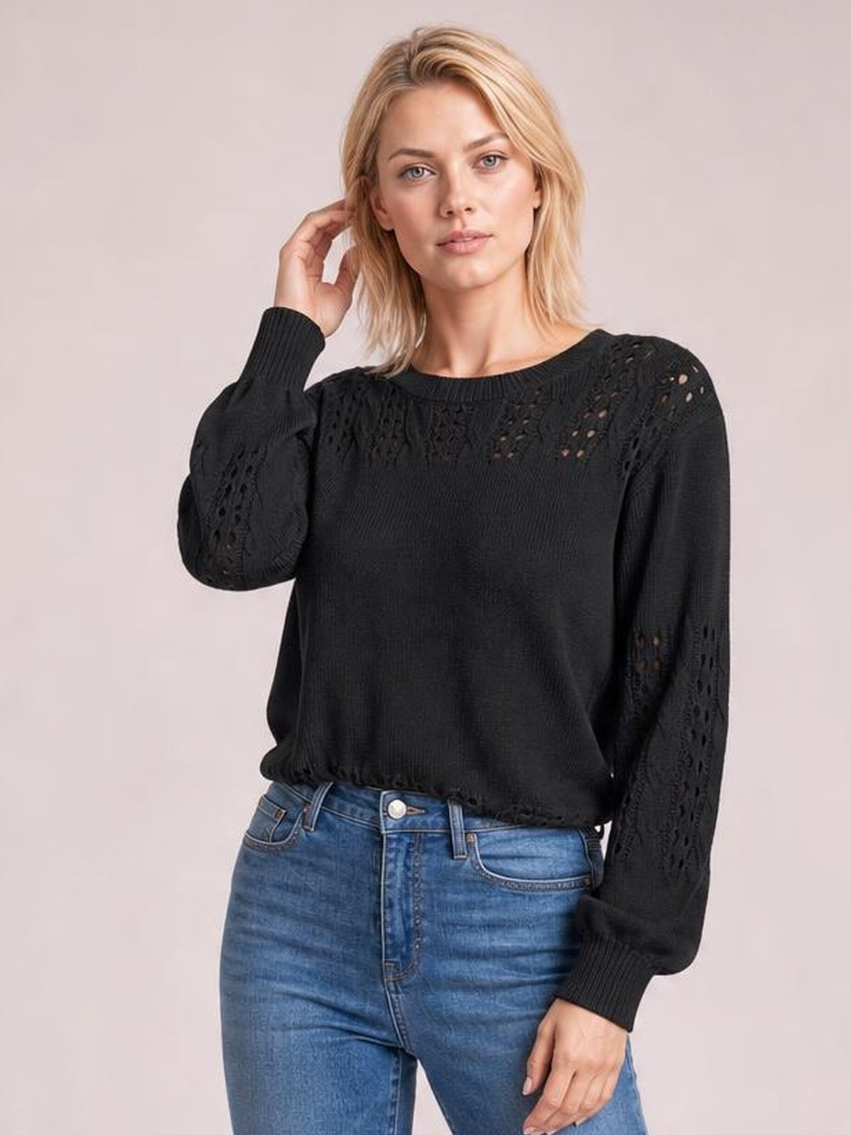 Model wears a black openwork round neck long sleeve sweater with a relaxed silhouette, ribbed cuffs and hem, posing with one hand near her hair.