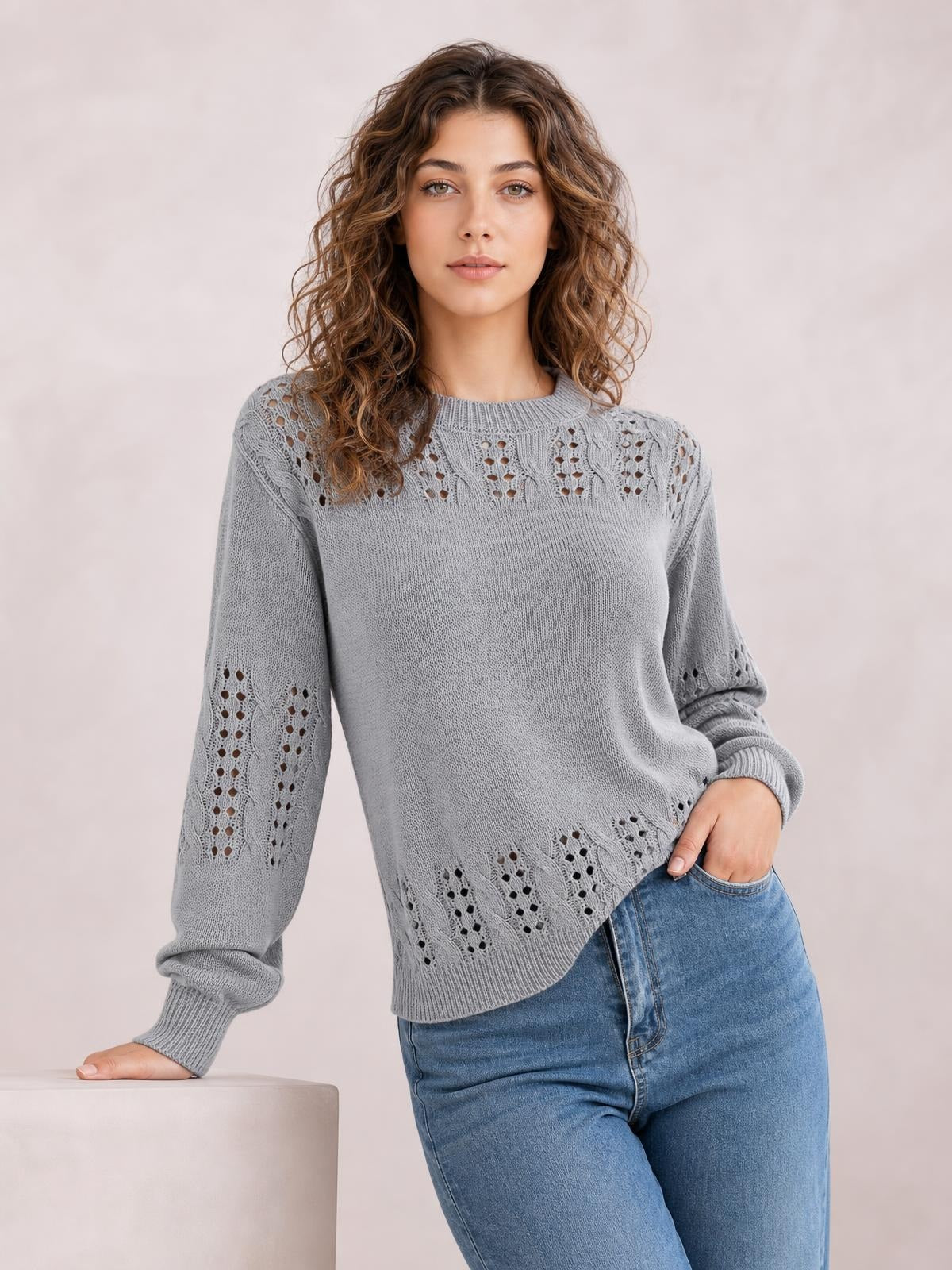 Model wearing a light gray openwork round neck long sleeve sweater with ribbed trim, relaxed silhouette, and patterned knit panels, posing with one hand in her jeans pocket.