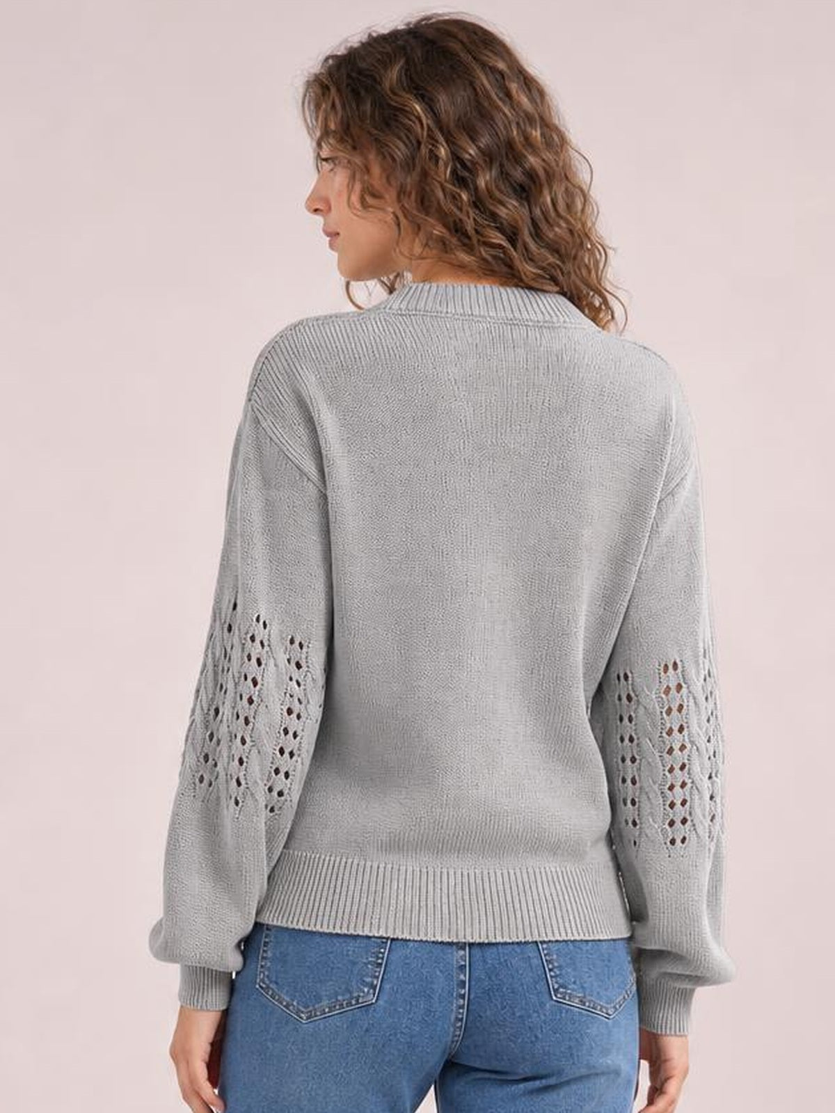 A model stands facing away in a light gray openwork round-neck sweater with long sleeves, decorative knit panels, ribbed cuffs and hem, and a relaxed silhouette.