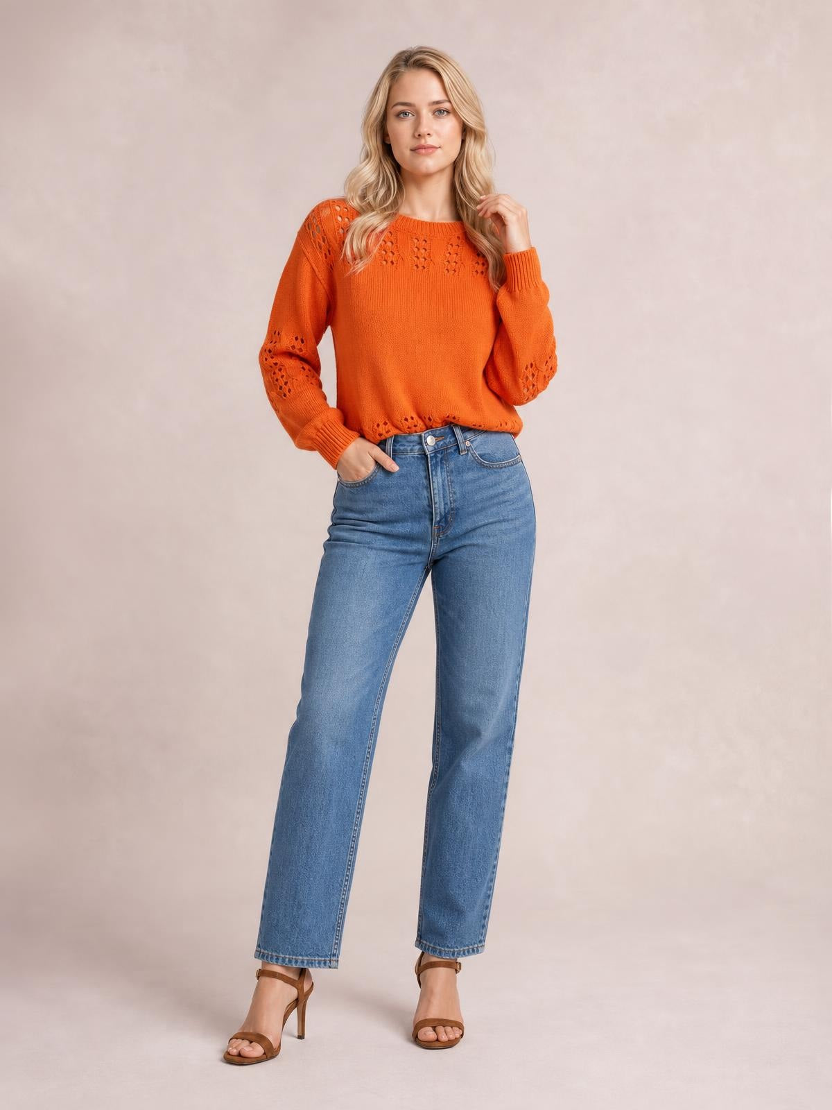 Model in a bright orange openwork round neck sweater with long sleeves, ribbed cuffs and hem, standing with one hand in her jeans pocket.