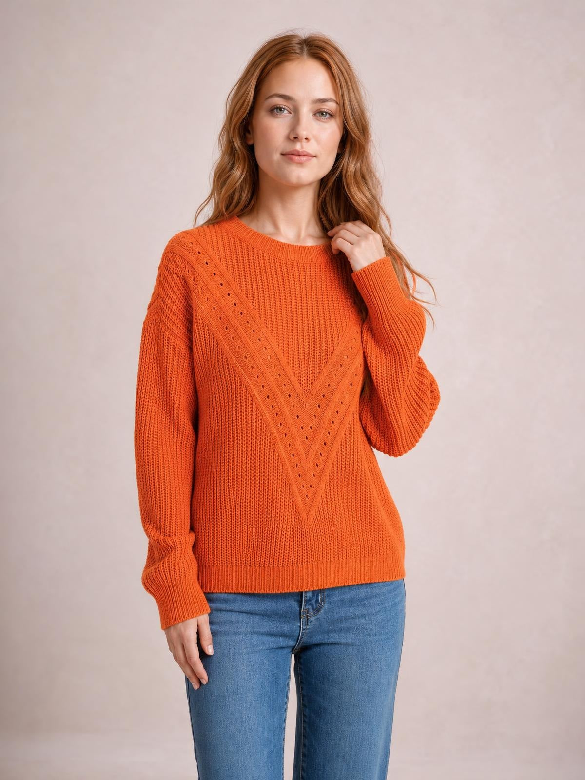 Model touches the neckline of an orange openwork round neck sweater with a relaxed silhouette, chevron knit detail, long sleeves, and ribbed cuffs and hem.