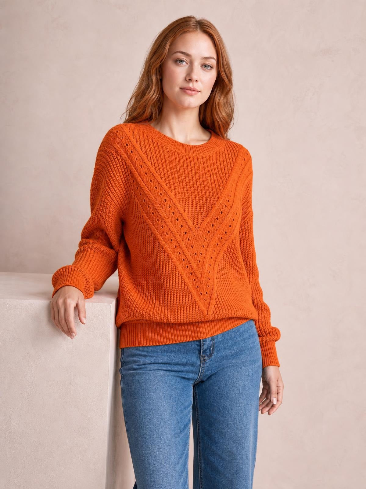 A model leans against a ledge wearing an orange openwork round neck sweater with a relaxed silhouette, ribbed cuffs and hem, and eyelet knit panels.