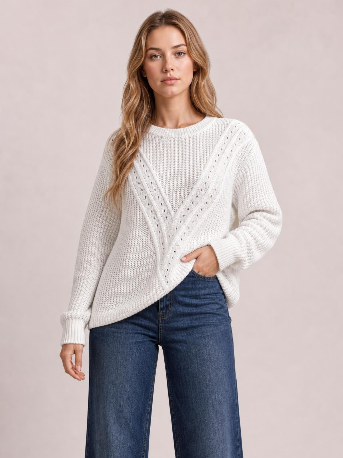 Model wearing a white openwork round neck sweater with a relaxed silhouette, ribbed cuffs and hem, and V-shaped knit detailing, posing with one hand in her jeans pocket.