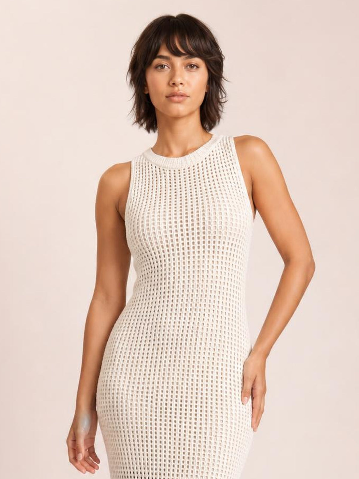 A model wears a cream semi-sheer openwork round-neck cover-up with a sleeveless design and fitted, body-skimming silhouette, standing with one hand on her hip.