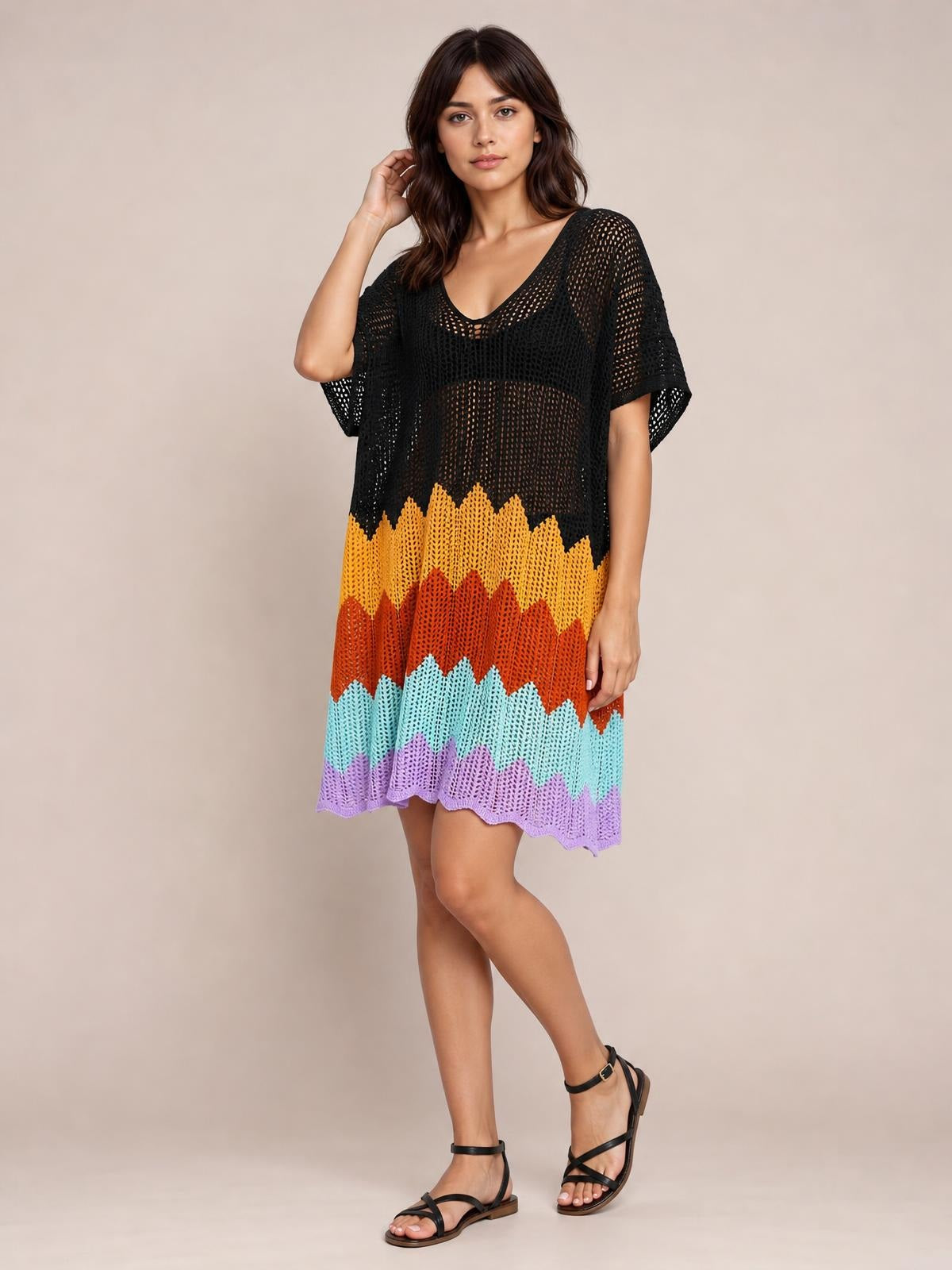 Model wears a loose openwork V-neck half-sleeve cover-up with a black bodice, colorful zigzag stripes, and a scalloped hem, posing with crossed legs.