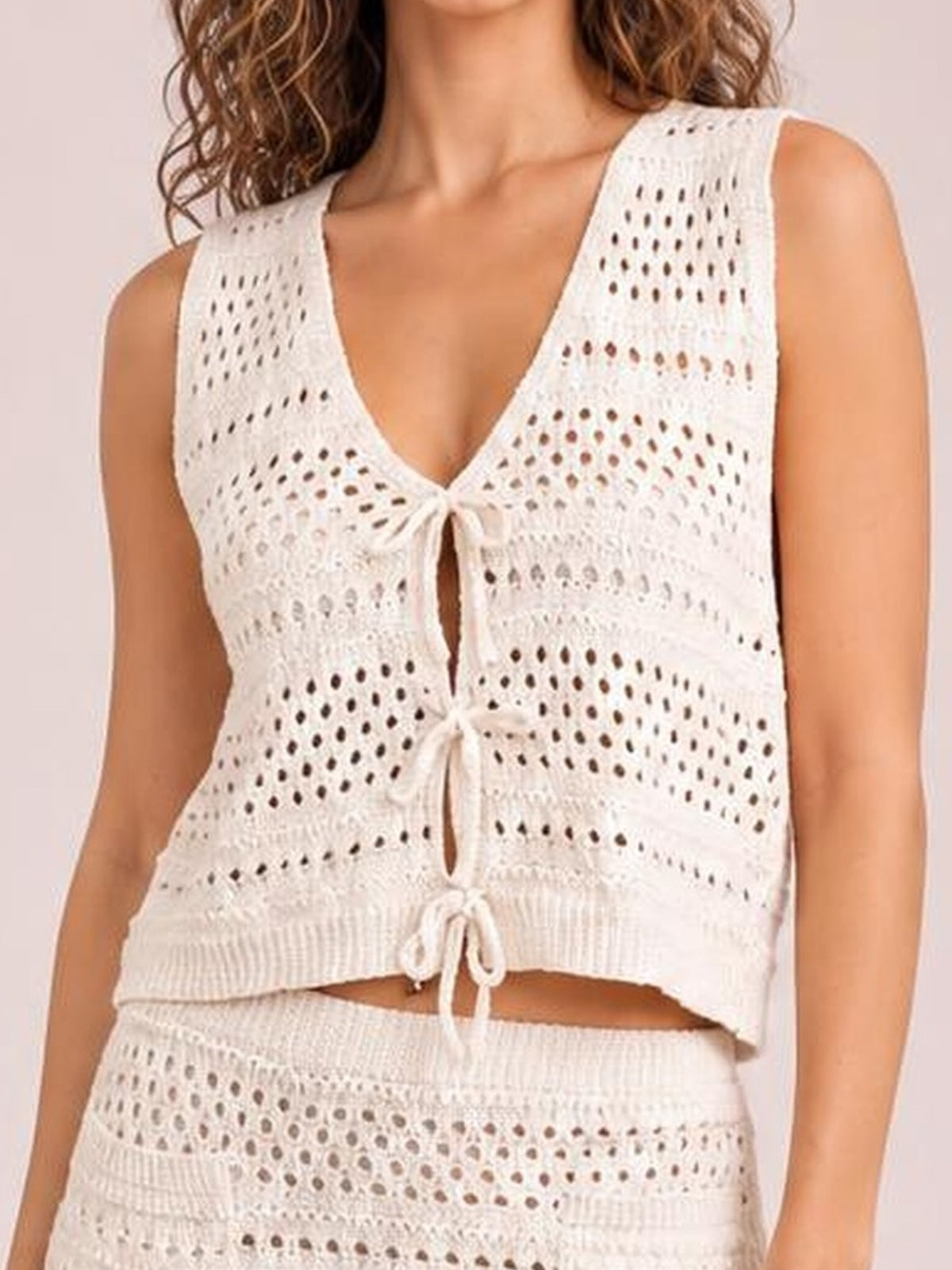 Model wears a white openwork knit cover-up set with a tied V-neck top, cropped silhouette, and matching patterned skirt.